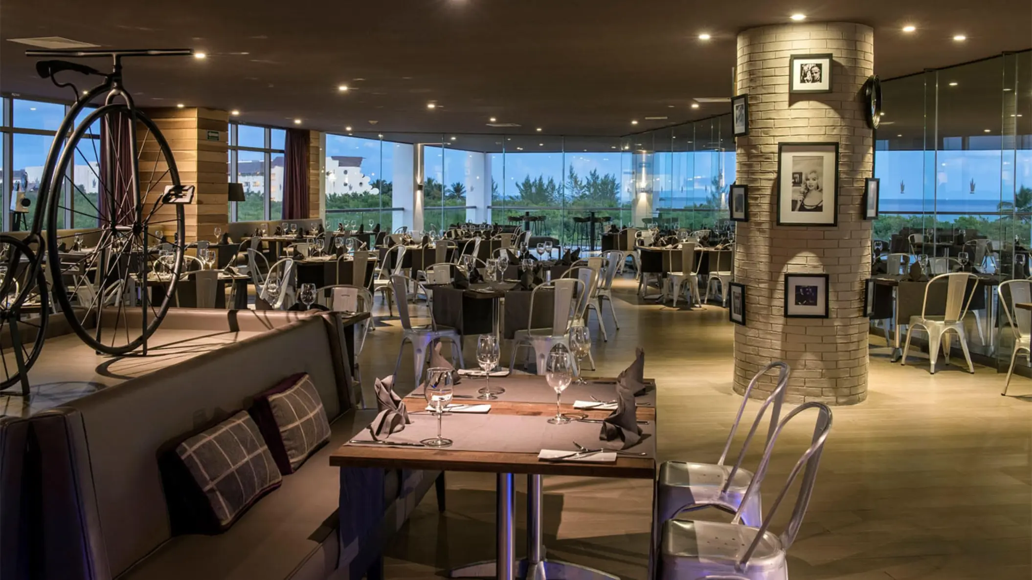 Interior of a hotel restaurant with modern decor, a large decorative bicycle, and floor-to-ceiling windows overlooking the ocean.