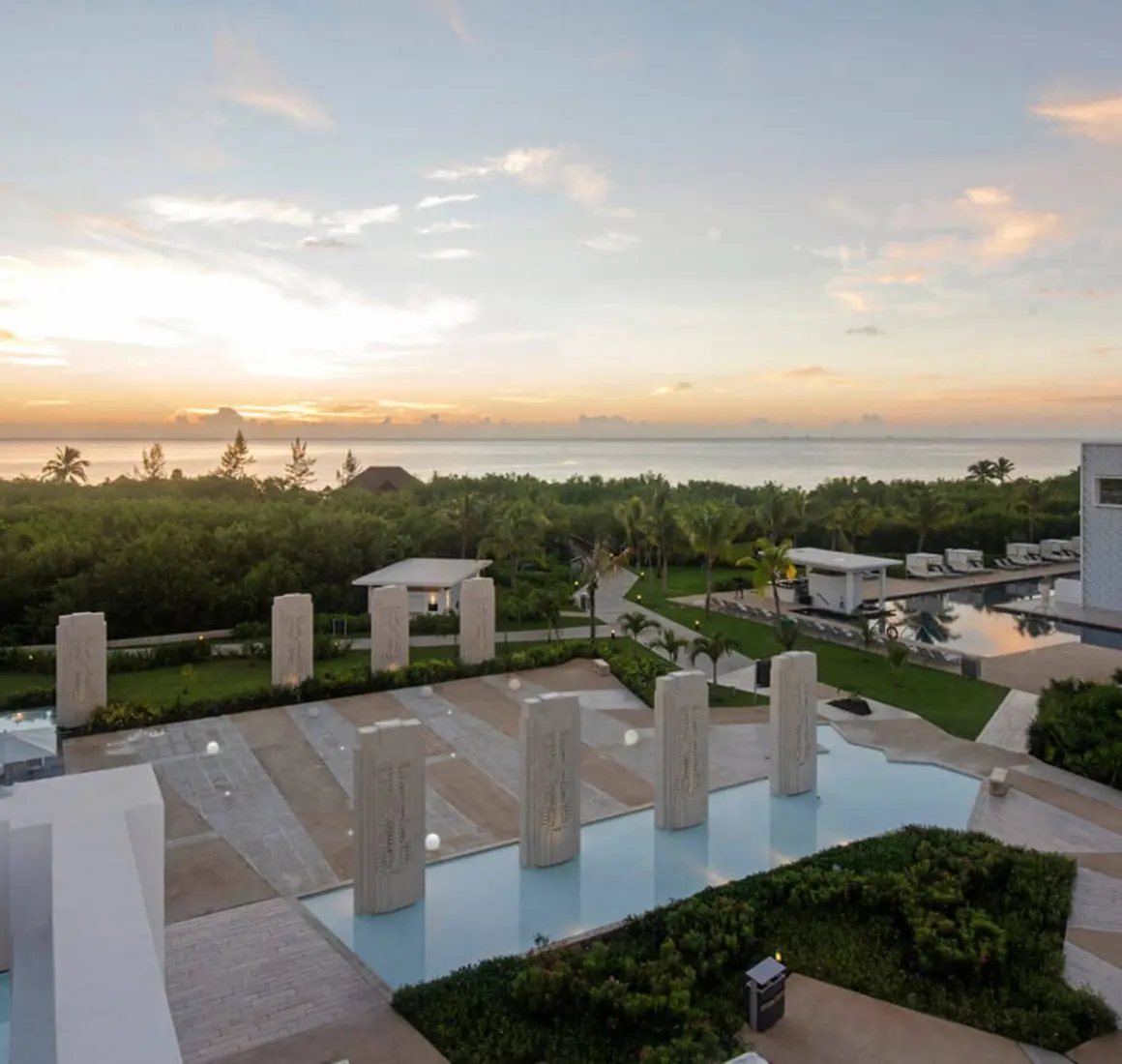 Sunset view from the rooftop, showing a decorative water feature, surrounding foliage, and the ocean in the distance.