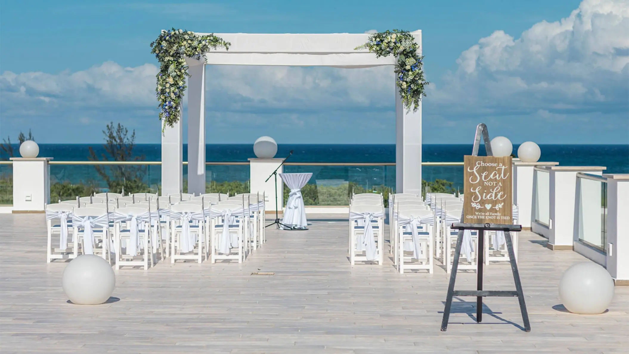 Rooftop wedding venue setup with a white pergola decorated with flowers, white chairs, and the ocean as the backdrop.