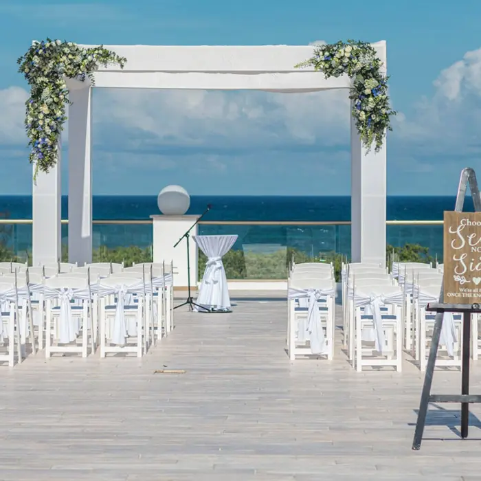 Rooftop wedding venue setup with a white pergola decorated with flowers, white chairs, and the ocean as the backdrop.