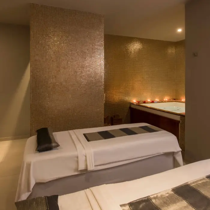 A relaxing spa treatment room with two massage tables, a jacuzzi area with candles, and a modern sink.