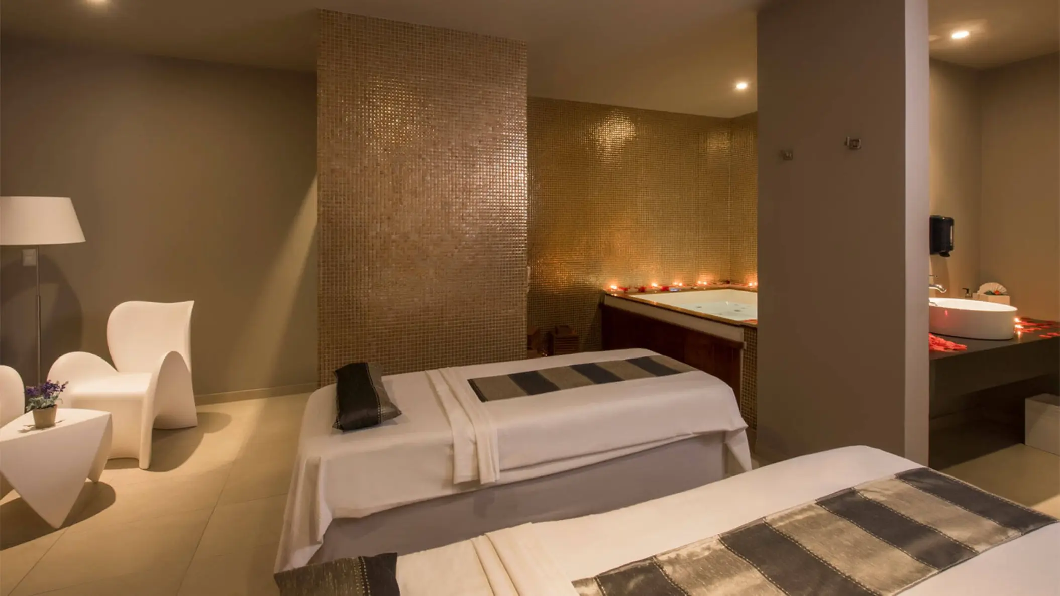 A relaxing spa treatment room with two massage tables, a jacuzzi area with candles, and a modern sink.