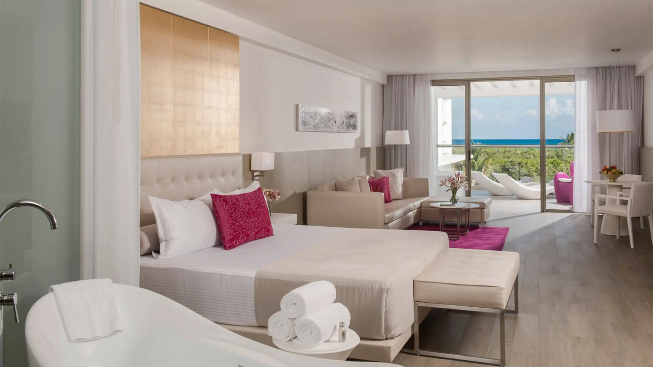 spacious hotel suite with a king-sized bed, sofa, dining area, and balcony overlooking the pool and ocean.