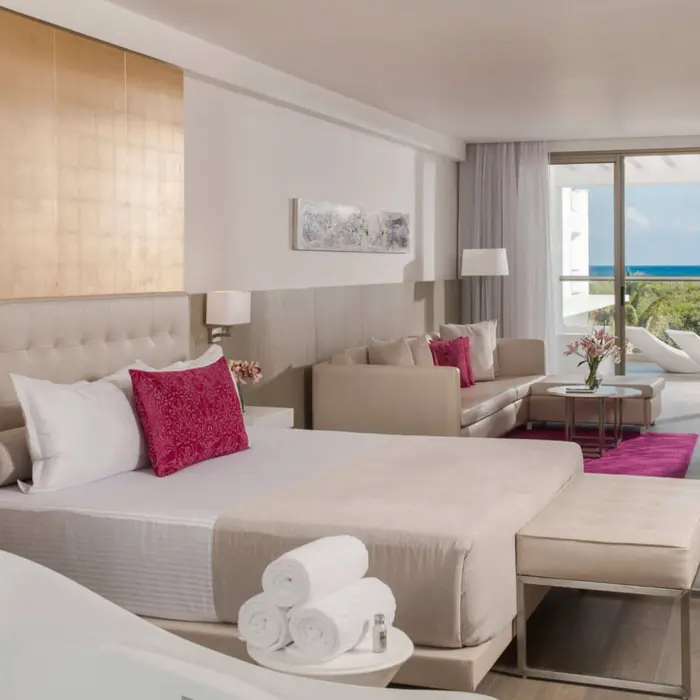 spacious hotel suite with a king-sized bed, sofa, dining area, and balcony overlooking the pool and ocean.