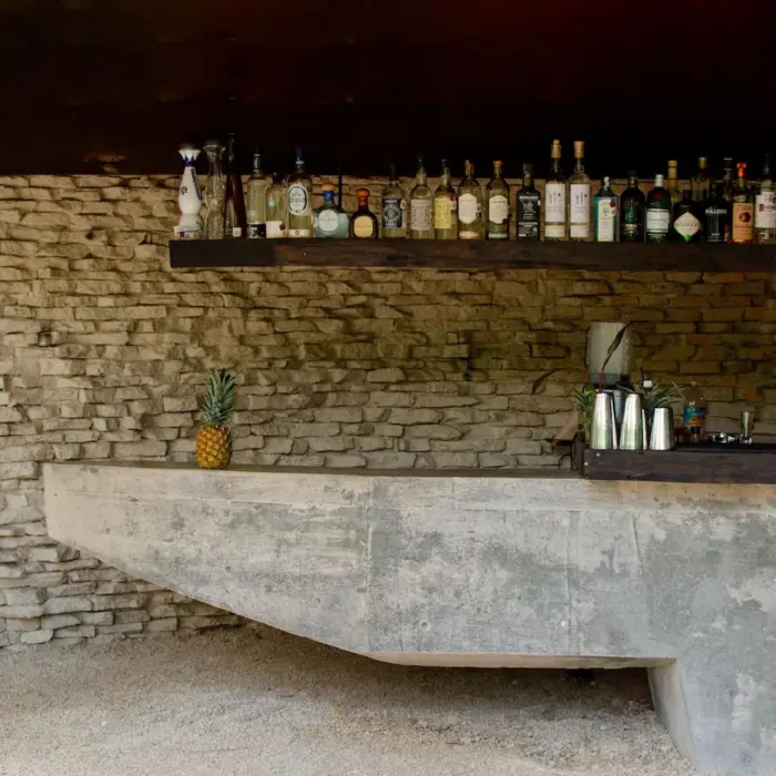 Rustic bar counter at Radhoo Tulum with warm lighting and artisanal bottles on wooden display shelves.