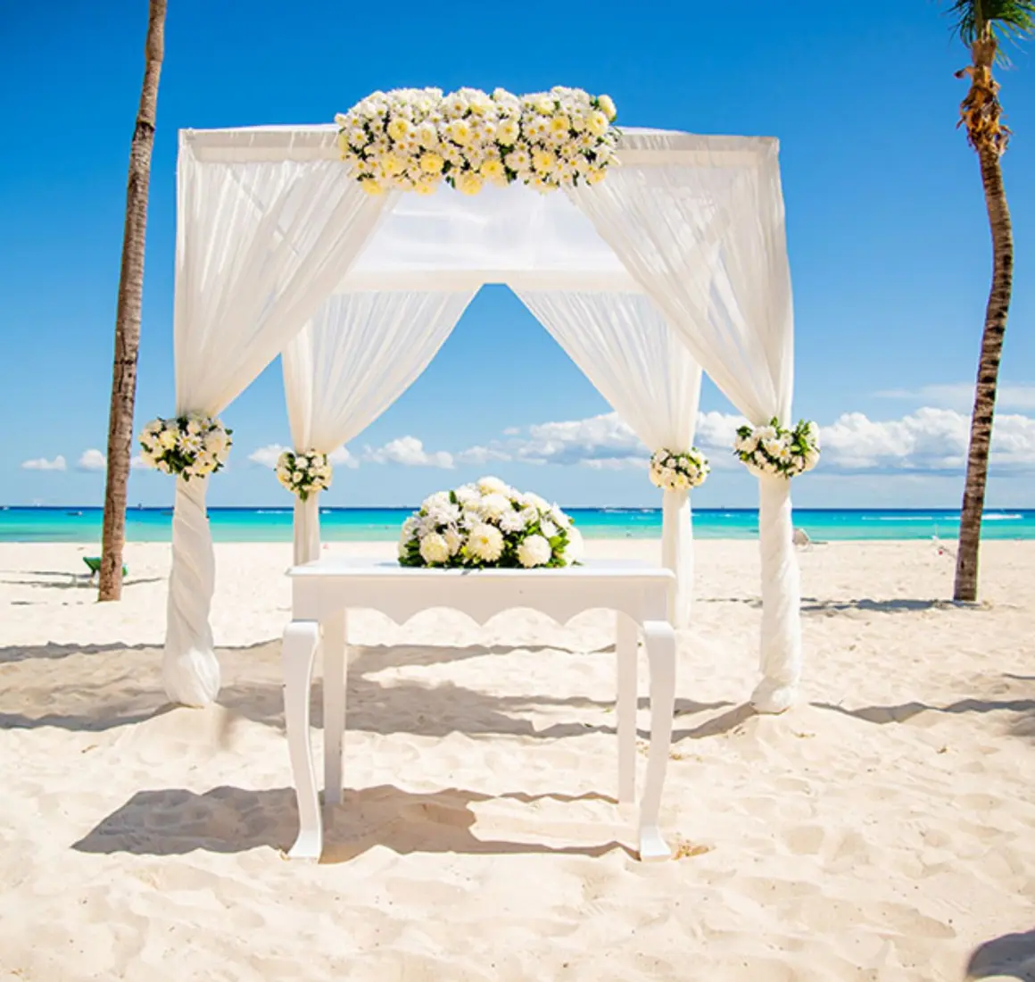 A white wedding gazebo on a pristine beach, with the turquoise ocean and palm trees in the background.