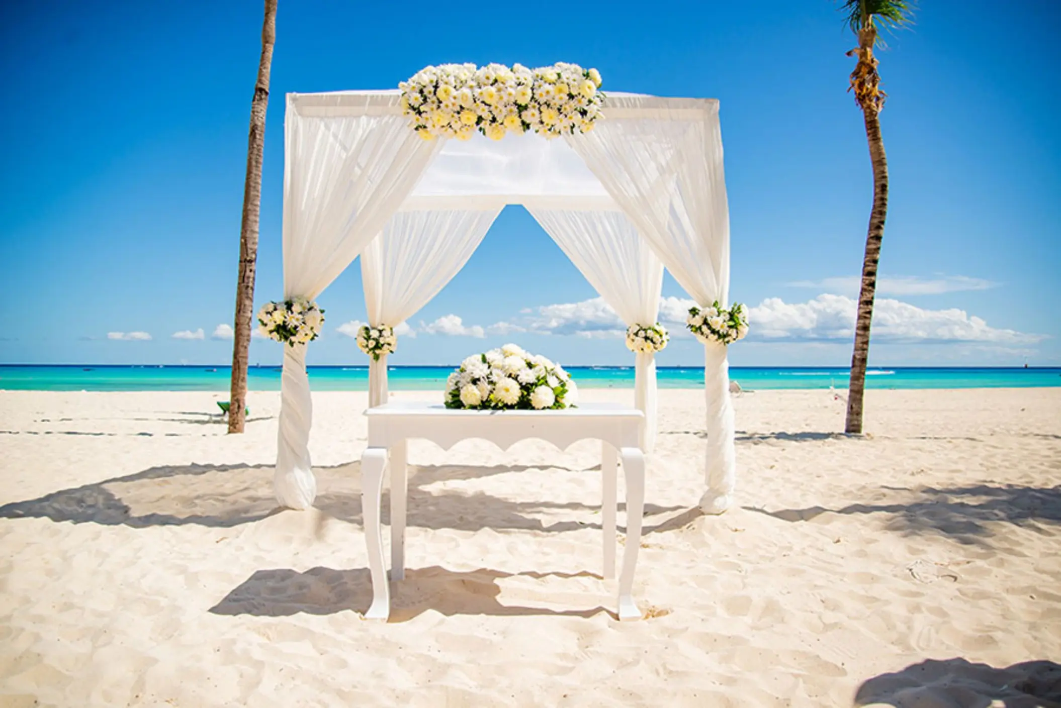 A white wedding gazebo on a pristine beach, with the turquoise ocean and palm trees in the background.