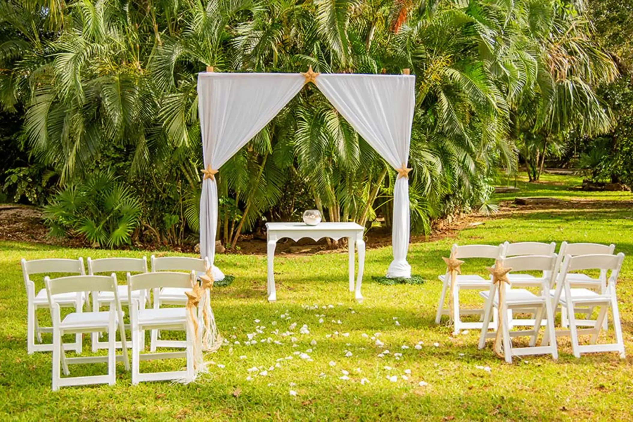 A beautiful garden wedding setup with a white gazebo, folding chairs, and scattered rose petals.