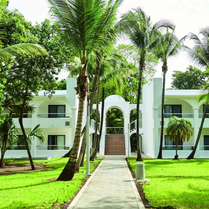 A walkway leading to a two-story hotel building, surrounded by lush green lawns and palm trees.