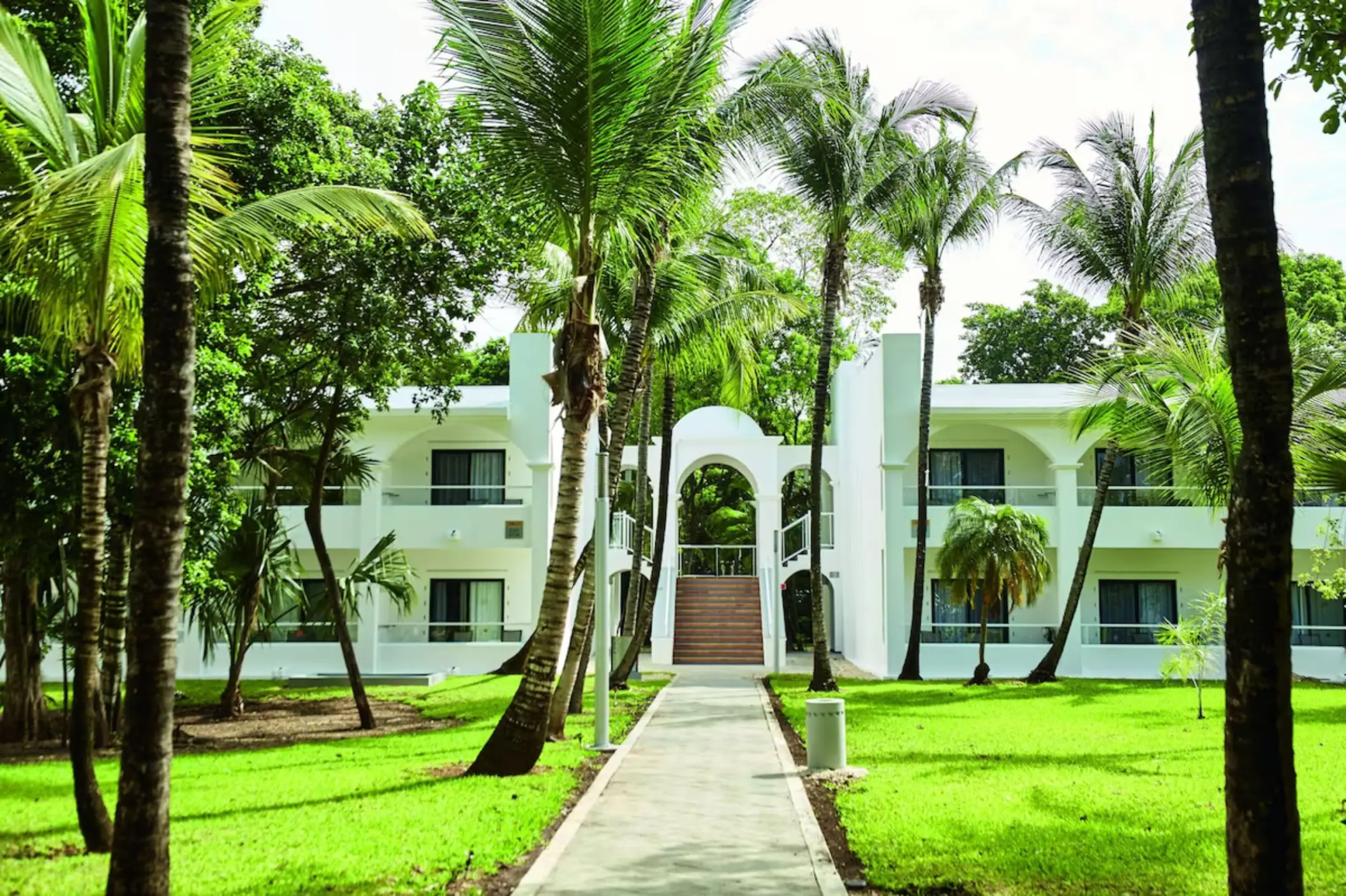 A walkway leading to a two-story hotel building, surrounded by lush green lawns and palm trees.