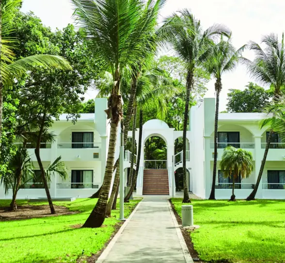 A walkway leading to a two-story hotel building, surrounded by lush green lawns and palm trees.