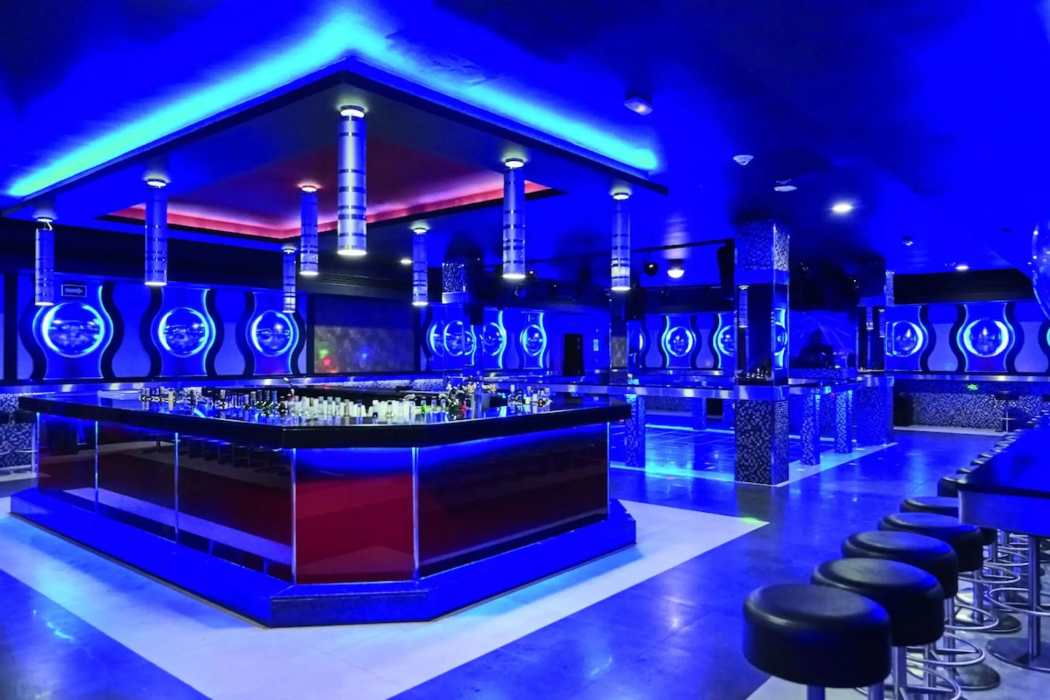 A modern, futuristic nightclub with blue lighting, a central bar, and lounge seating.