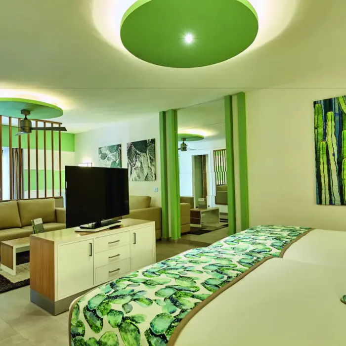 A spacious and bright suite with a bed and a separate living area with green accents and cactus art.