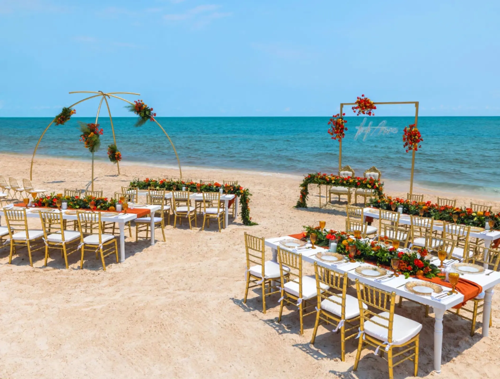 Elegant beach wedding setup with gold chairs, floral arch, and ocean backdrop under a bright blue sky.