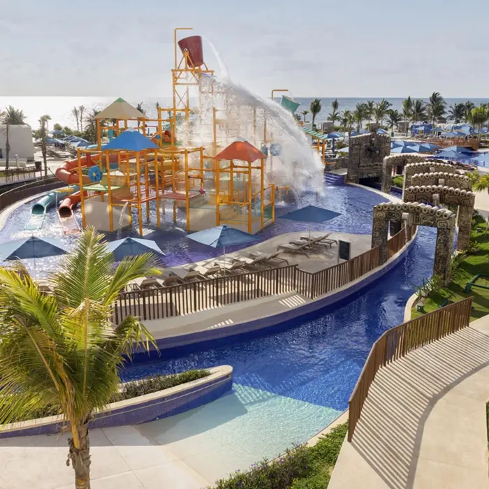 Outdoor water park with splash buckets, water slides, and climbing structures surrounded by lounge chairs and umbrellas.