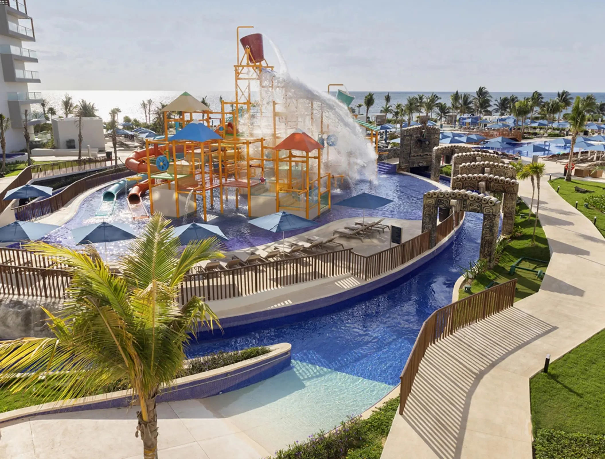 Outdoor water park with splash buckets, water slides, and climbing structures surrounded by lounge chairs and umbrellas.