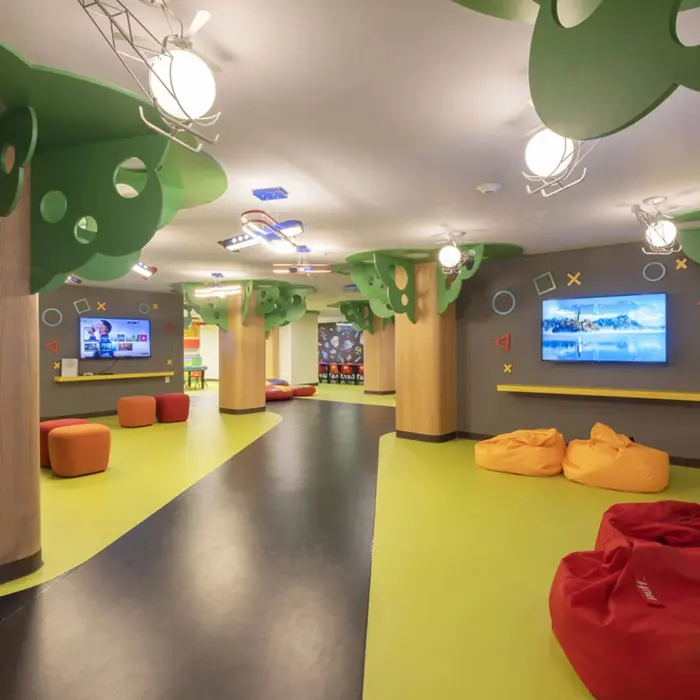 Colorful kids club with bean bags, wall-mounted TVs, tree-themed columns, and interactive play areas.