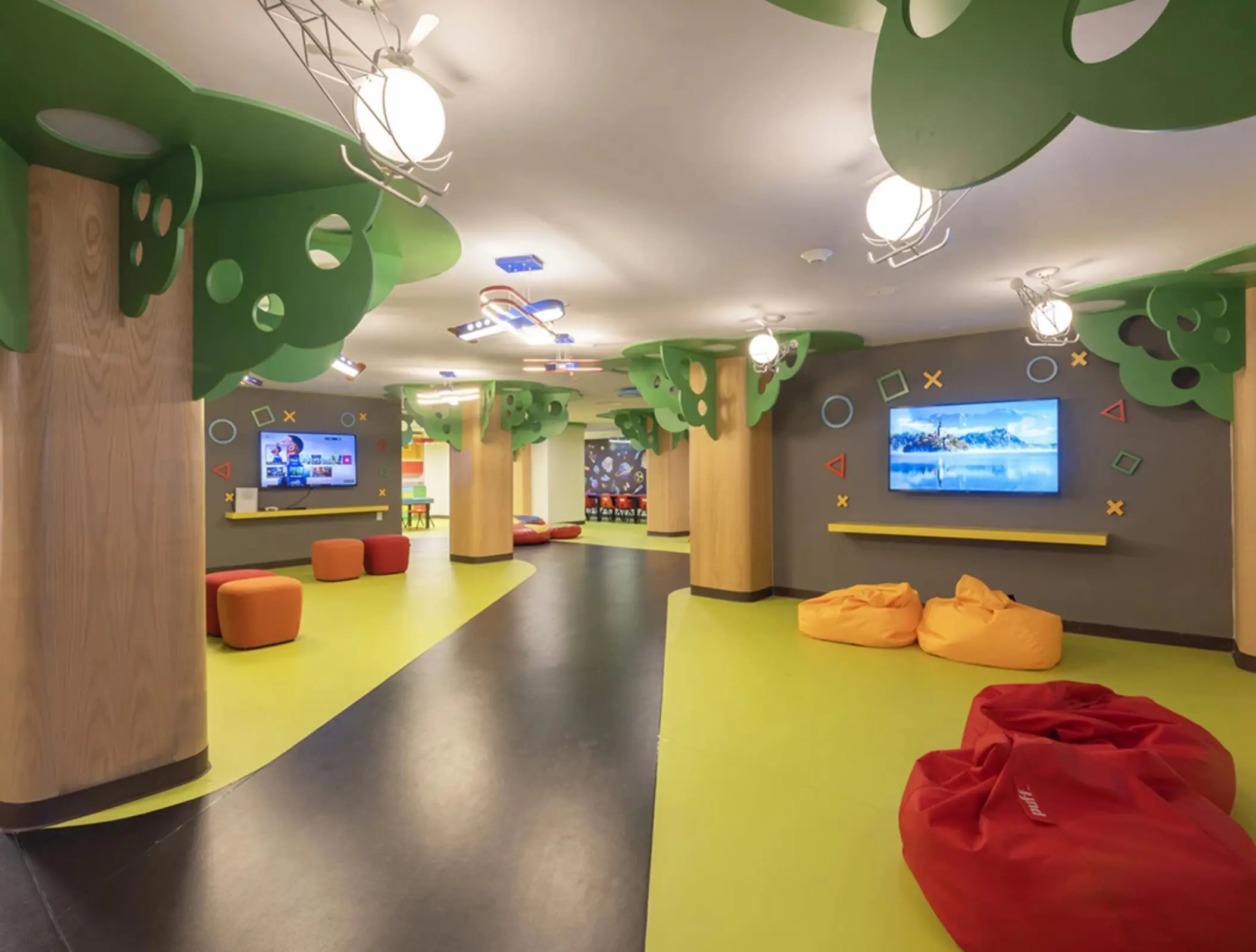 Colorful kids club with bean bags, wall-mounted TVs, tree-themed columns, and interactive play areas.
