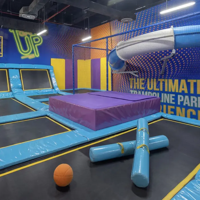 Indoor trampoline park with angled trampolines, padded obstacles, and a twisty slide against a vibrant wall mural.