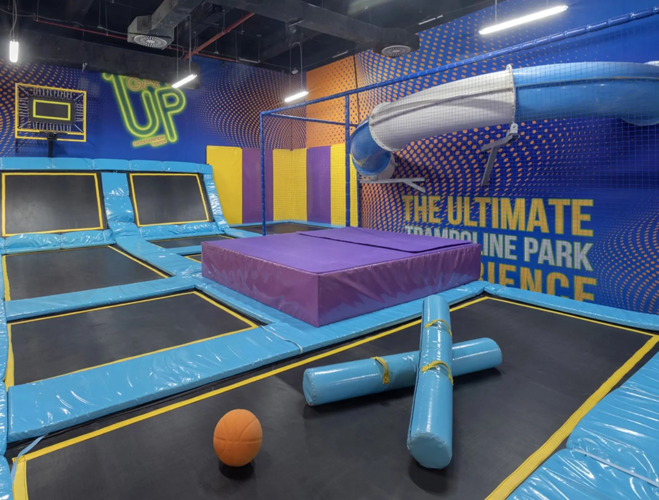 Indoor trampoline park with angled trampolines, padded obstacles, and a twisty slide against a vibrant wall mural.