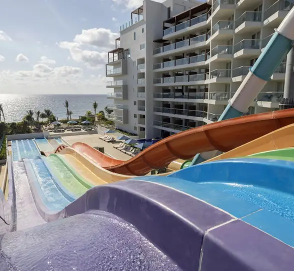 Colorful water slides with ocean views, pool loungers, and adjacent beachfront resort buildings under sunny skies.