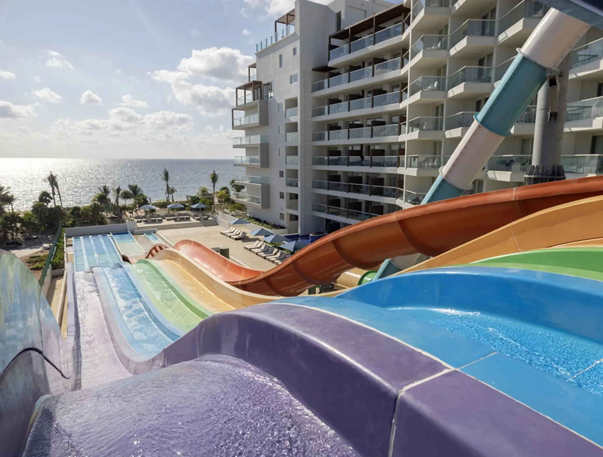 Colorful water slides with ocean views, pool loungers, and adjacent beachfront resort buildings under sunny skies.