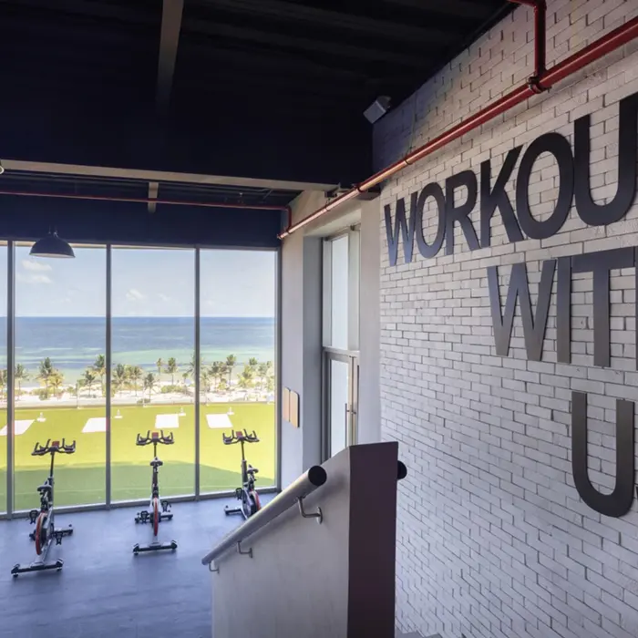 Ocean-view gym space with stationary bikes and “Workout With Us' signage, overlooking a lawn and beach area.