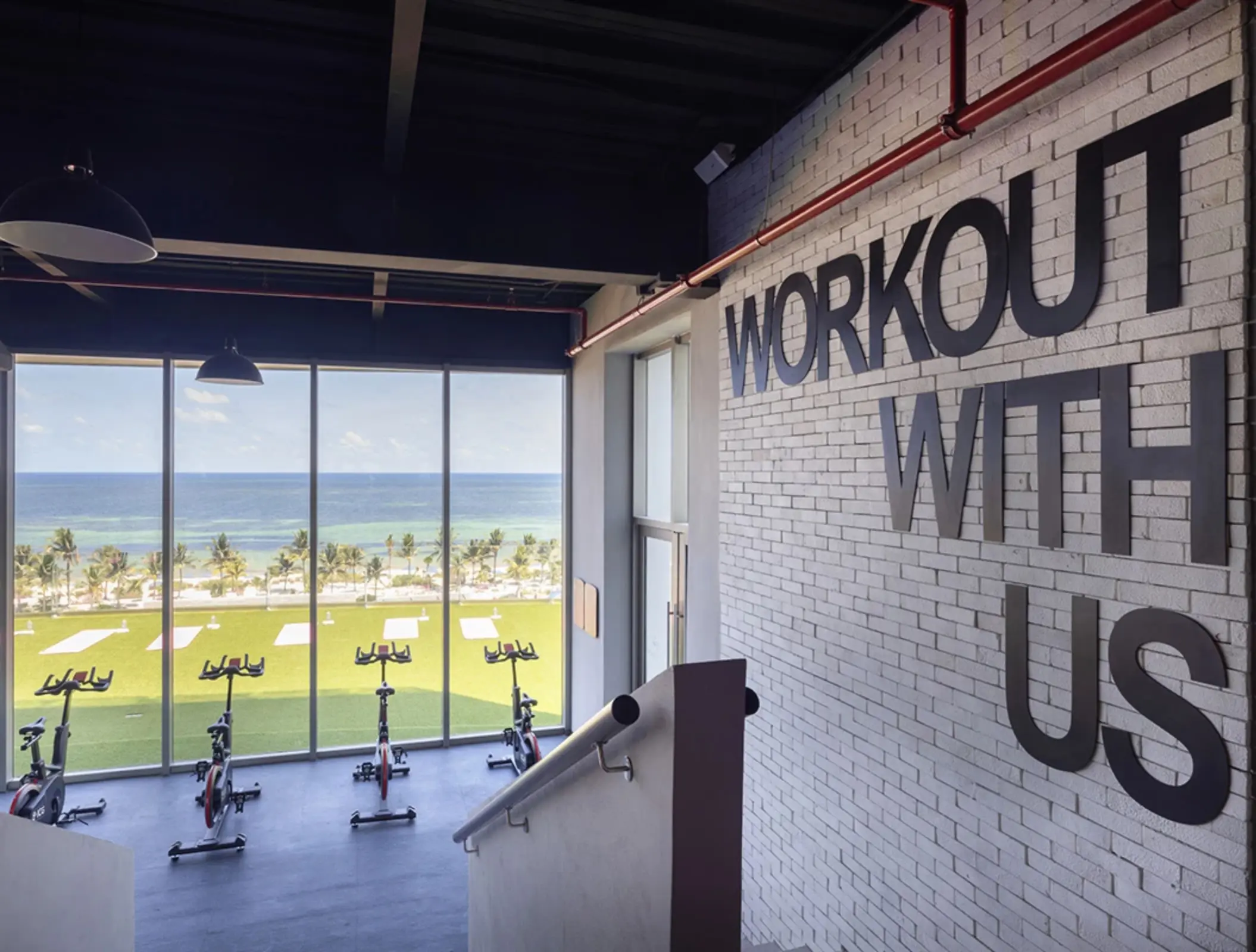 Ocean-view gym space with stationary bikes and “Workout With Us' signage, overlooking a lawn and beach area.