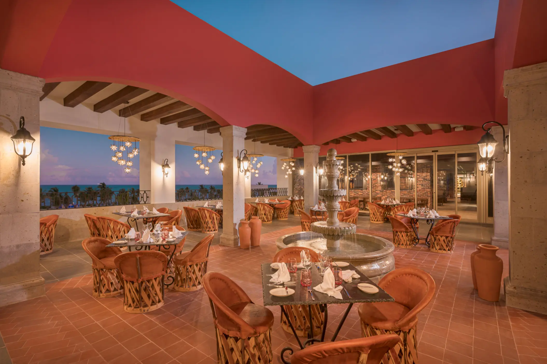Open-air terrace restaurant with terracotta floors, Mexican decor, and ocean views at sunset.