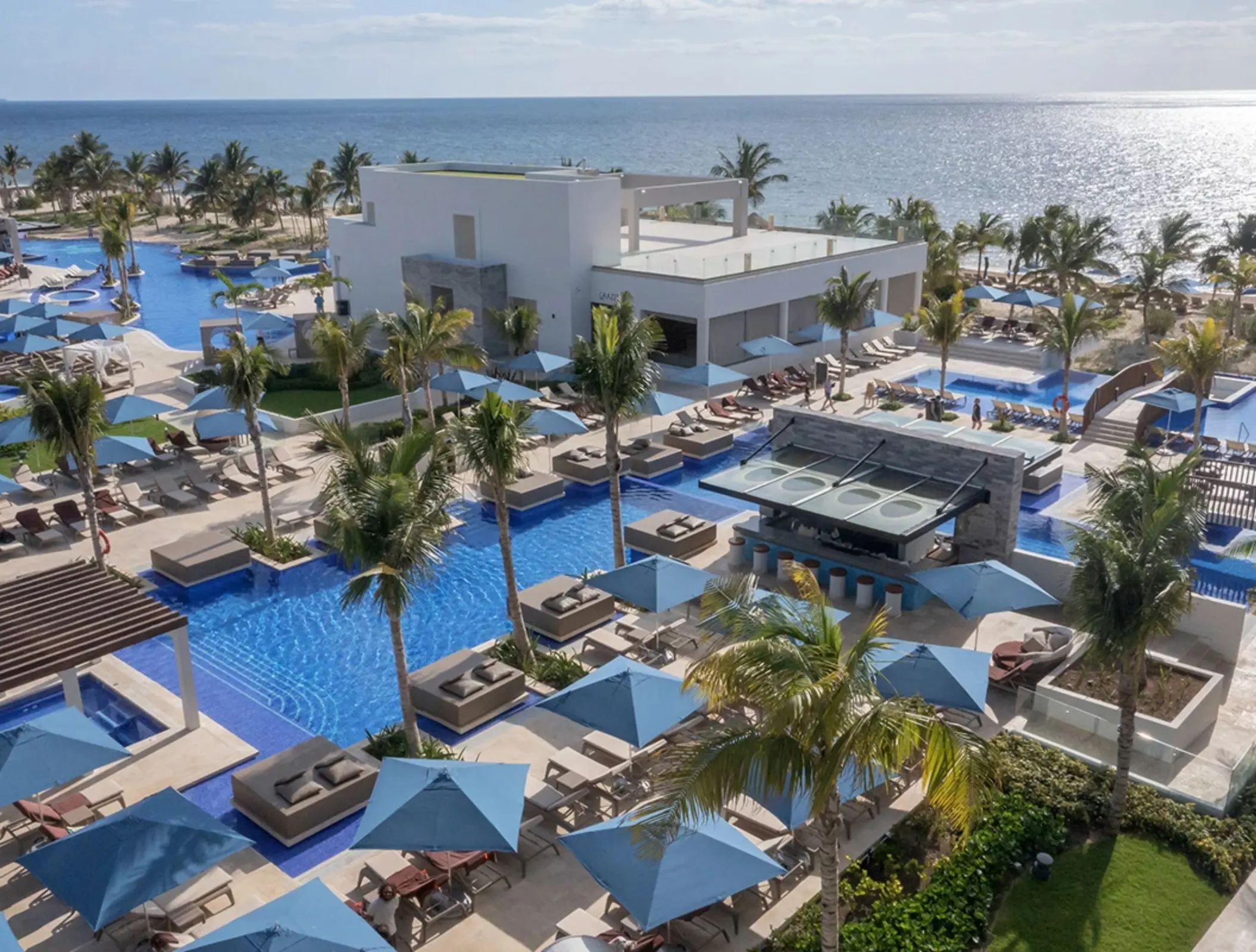 Overhead view of resort pools surrounded by palm trees, sunbeds, and a stylish swim-up bar by the beach.