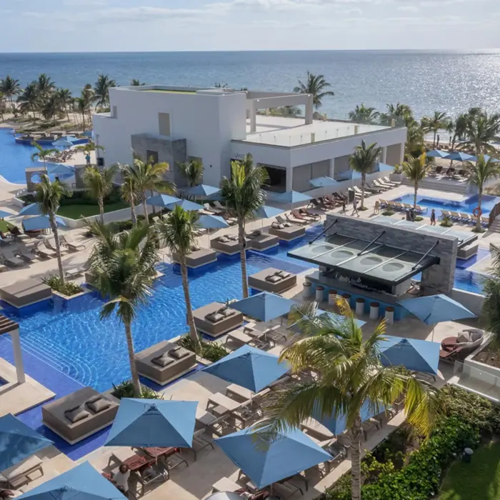 Overhead view of resort pools surrounded by palm trees, sunbeds, and a stylish swim-up bar by the beach.