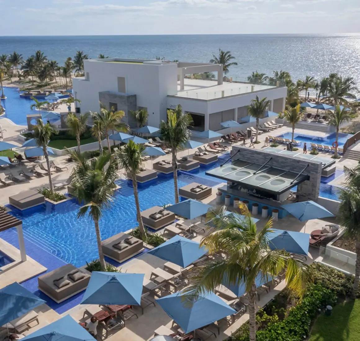 Overhead view of resort pools surrounded by palm trees, sunbeds, and a stylish swim-up bar by the beach.