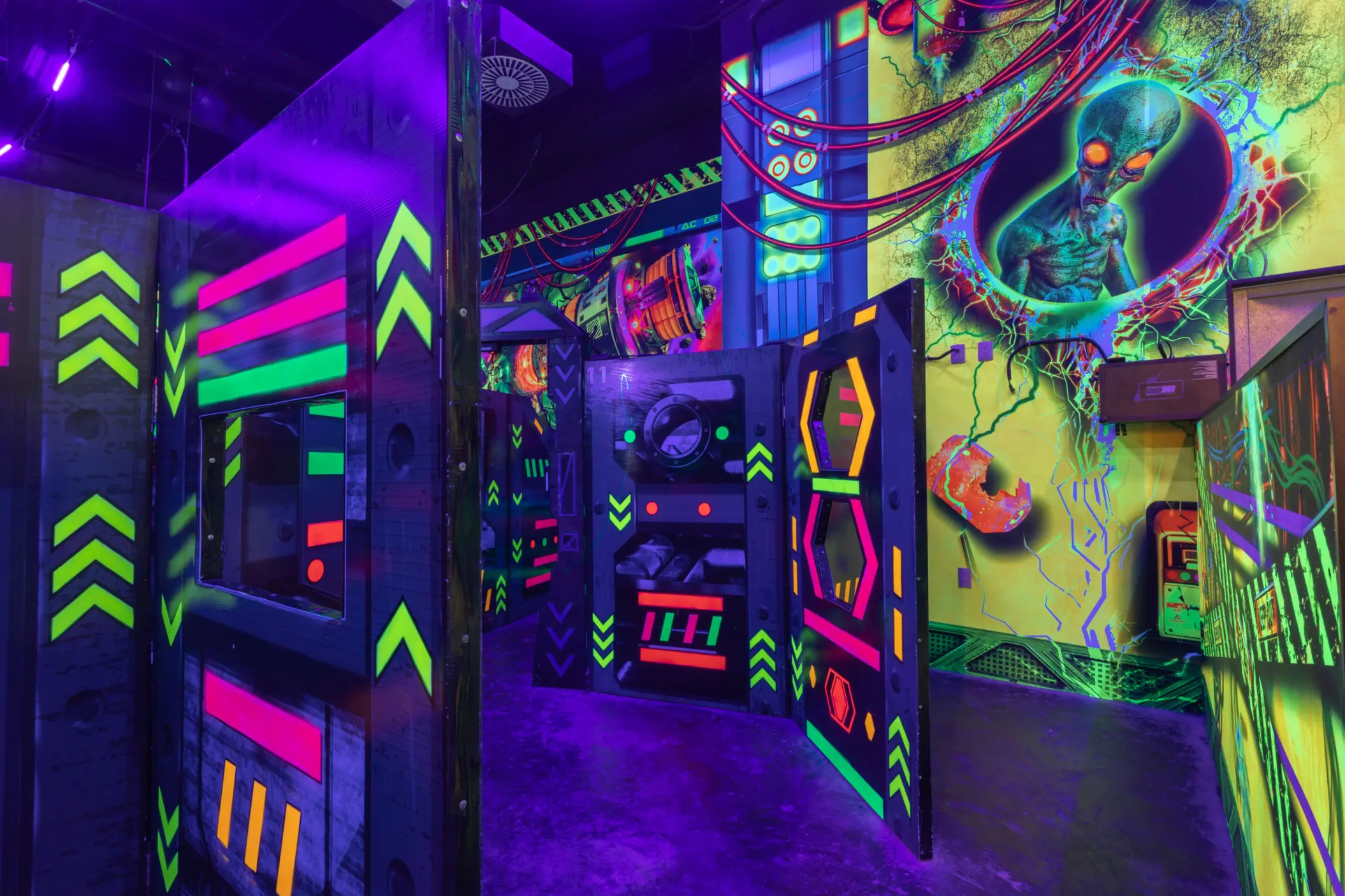 Neon-lit laser tag arena with alien-themed wall art and vibrant obstacles under blacklight for energetic play.