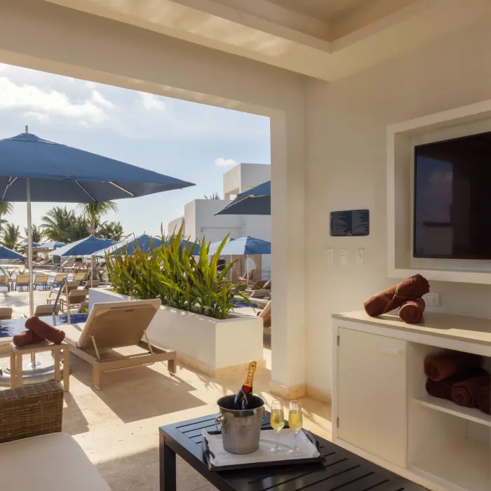 Private cabana with TV and champagne overlooking sun loungers and umbrellas beside a bright resort pool.