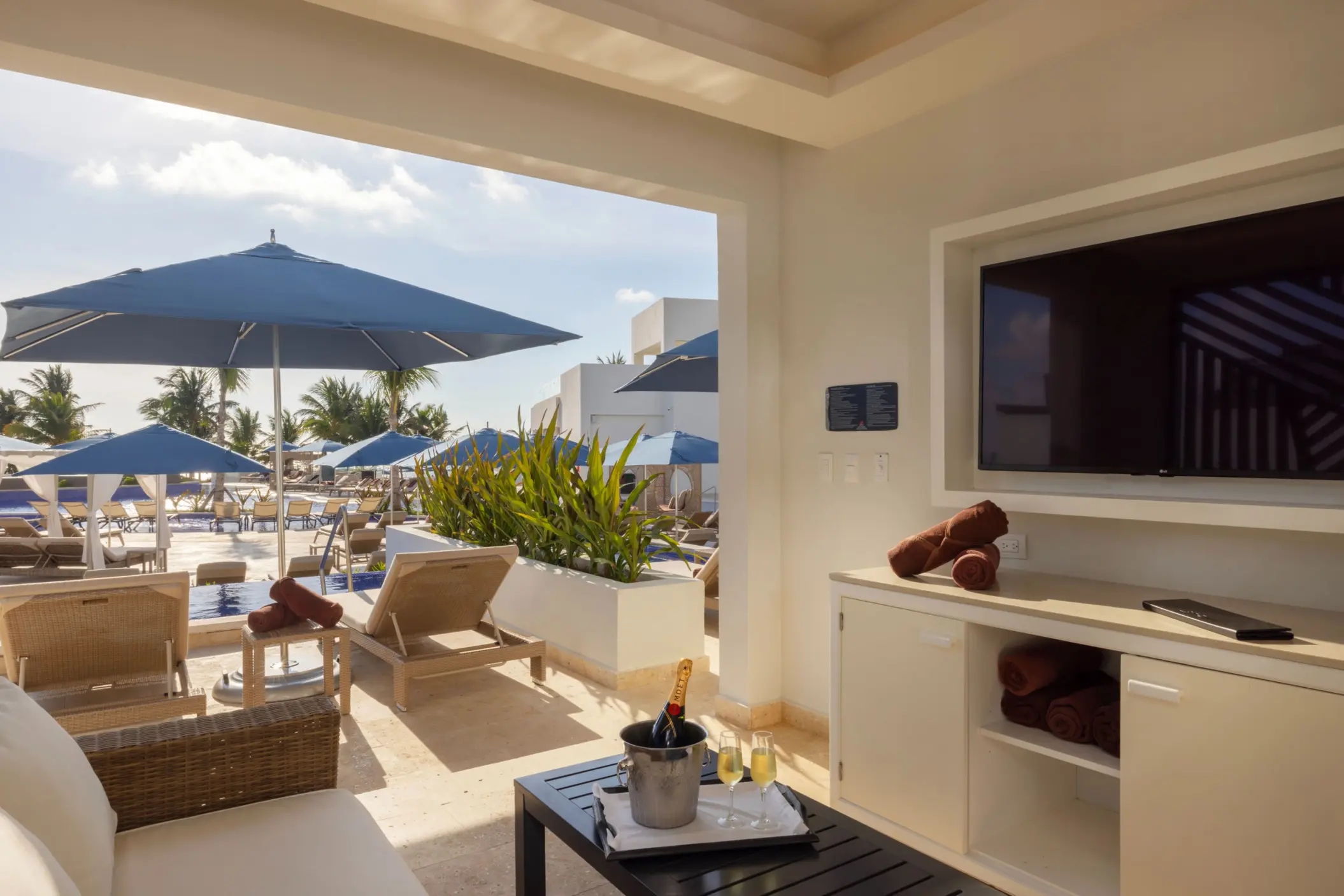 Private cabana with TV and champagne overlooking sun loungers and umbrellas beside a bright resort pool.