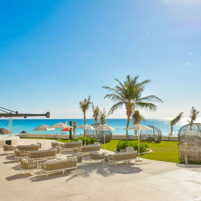 An outdoor lounge area with chairs, tables, and palm trees, located on a patio next to the beach.