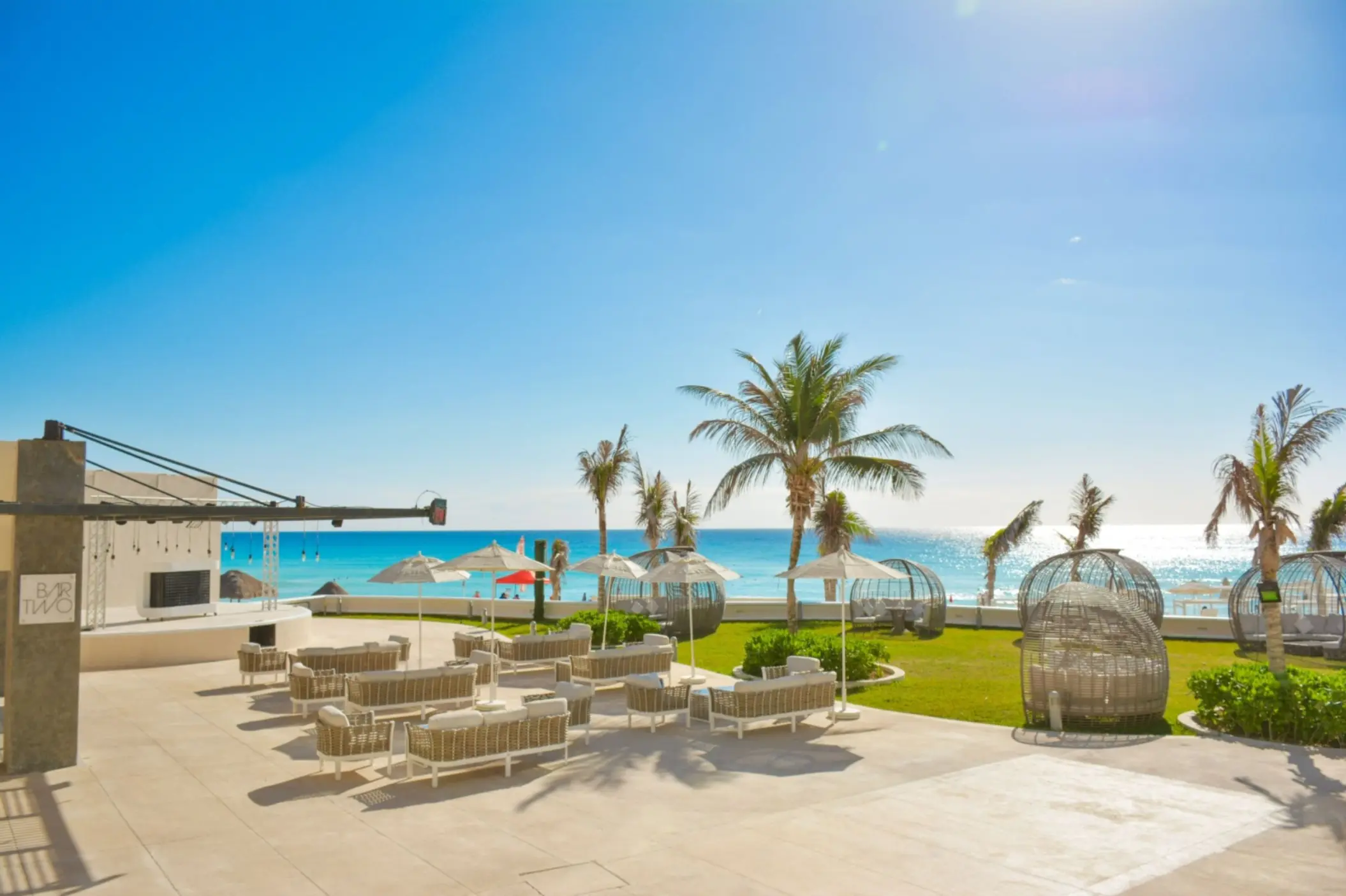 An outdoor lounge area with chairs, tables, and palm trees, located on a patio next to the beach.