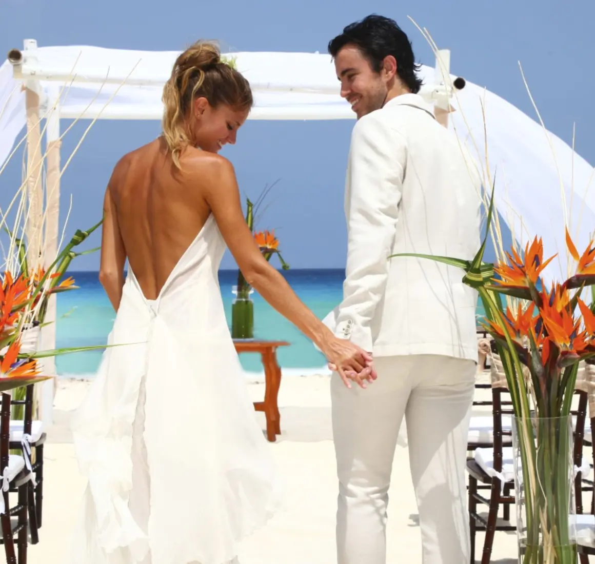 A bride and groom holding hands and walking on a white sand beach toward a wedding arch.