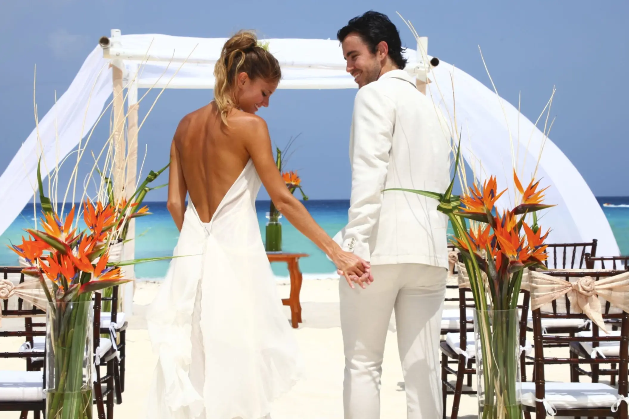 A bride and groom holding hands and walking on a white sand beach toward a wedding arch.