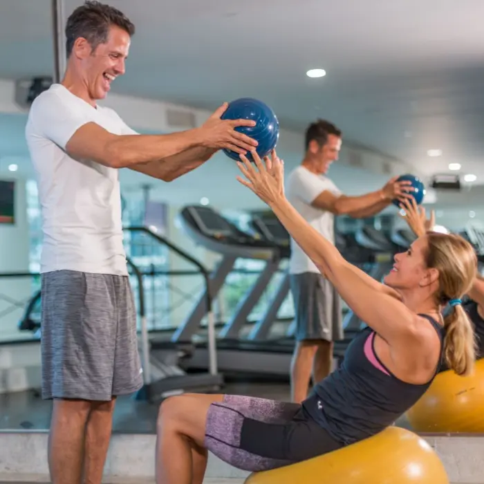 A happy couple working out in a gym, with the man helping the woman with a medicine ball crunch.
