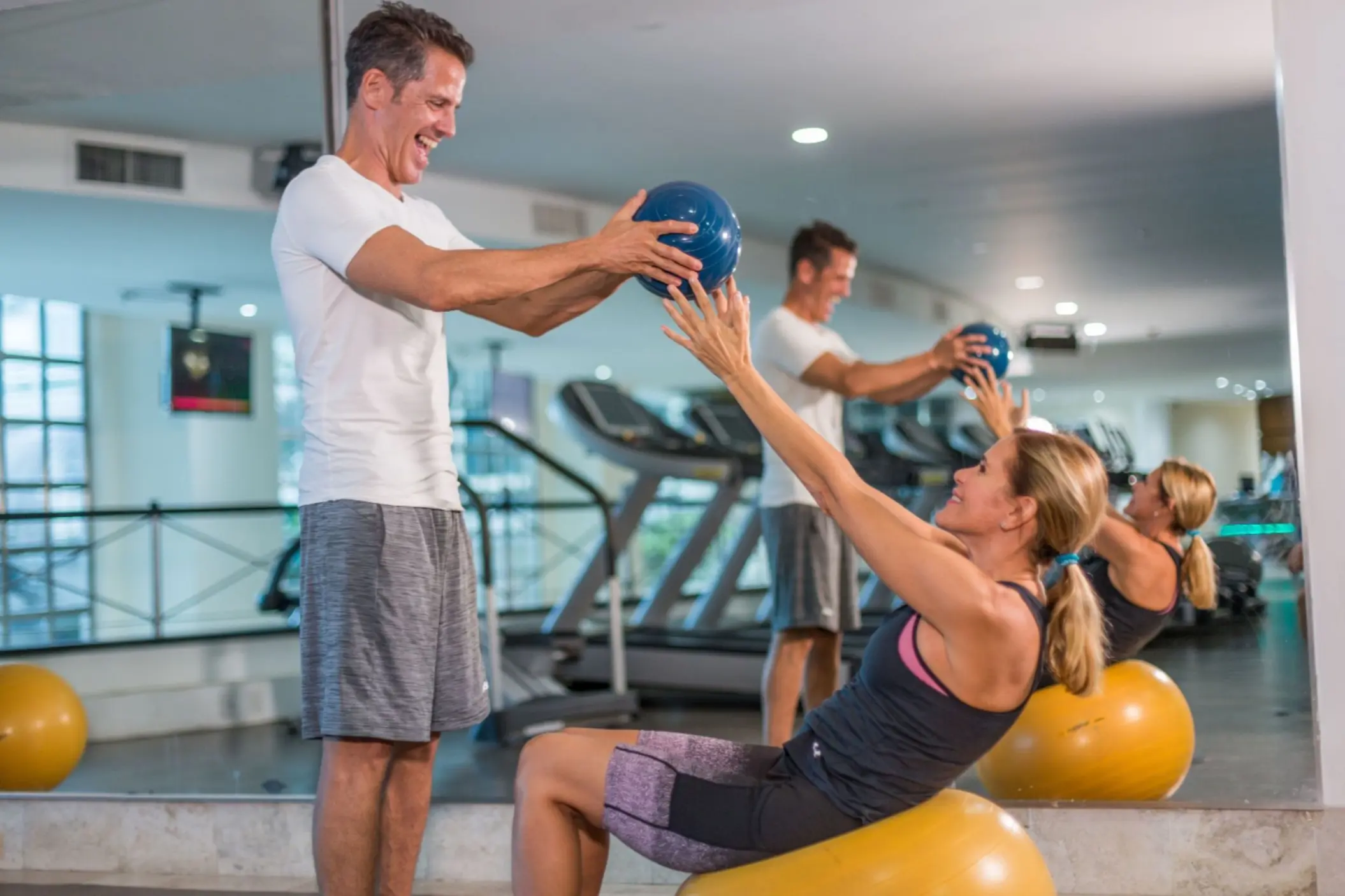 A happy couple working out in a gym, with the man helping the woman with a medicine ball crunch.