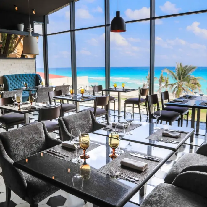 A modern restaurant dining room with floor-to-ceiling windows and tables overlooking the beach and ocean.