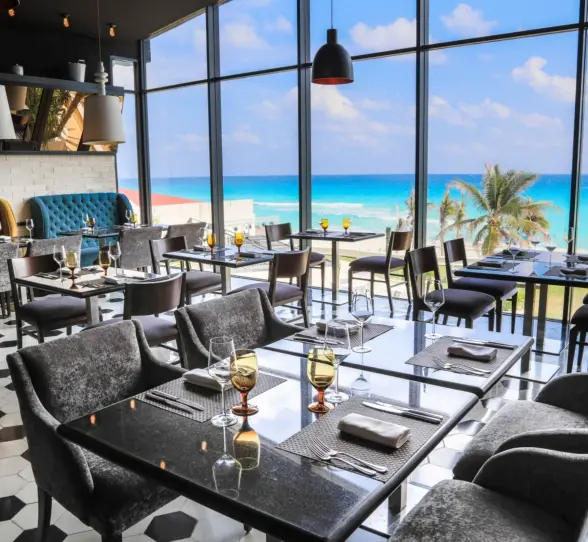 A modern restaurant dining room with floor-to-ceiling windows and tables overlooking the beach and ocean.