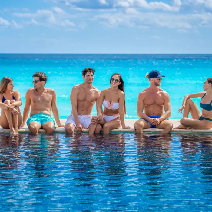A group of friends, including two couples, sitting on the edge of a hotel pool with a clear view of the ocean.