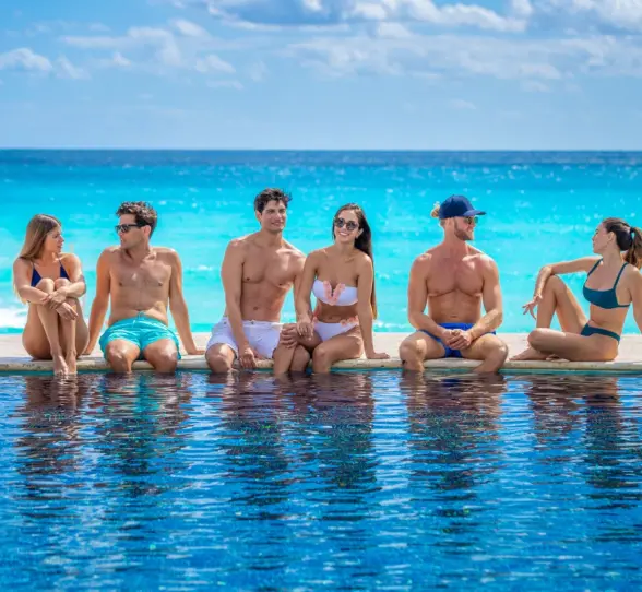 A group of friends, including two couples, sitting on the edge of a hotel pool with a clear view of the ocean.