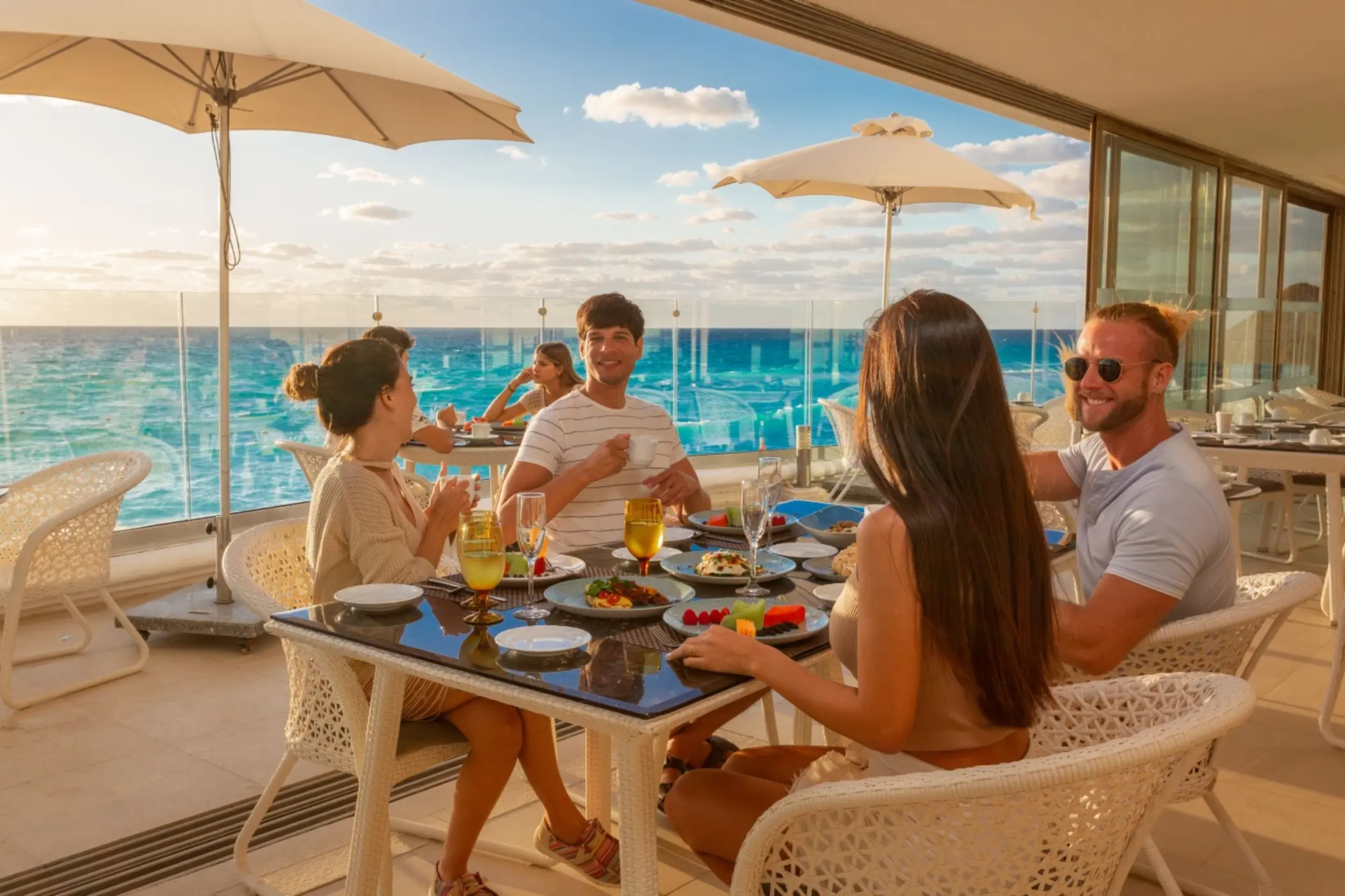 Guests enjoying an outdoor meal at a restaurant with white furniture and a stunning ocean view at sunset.