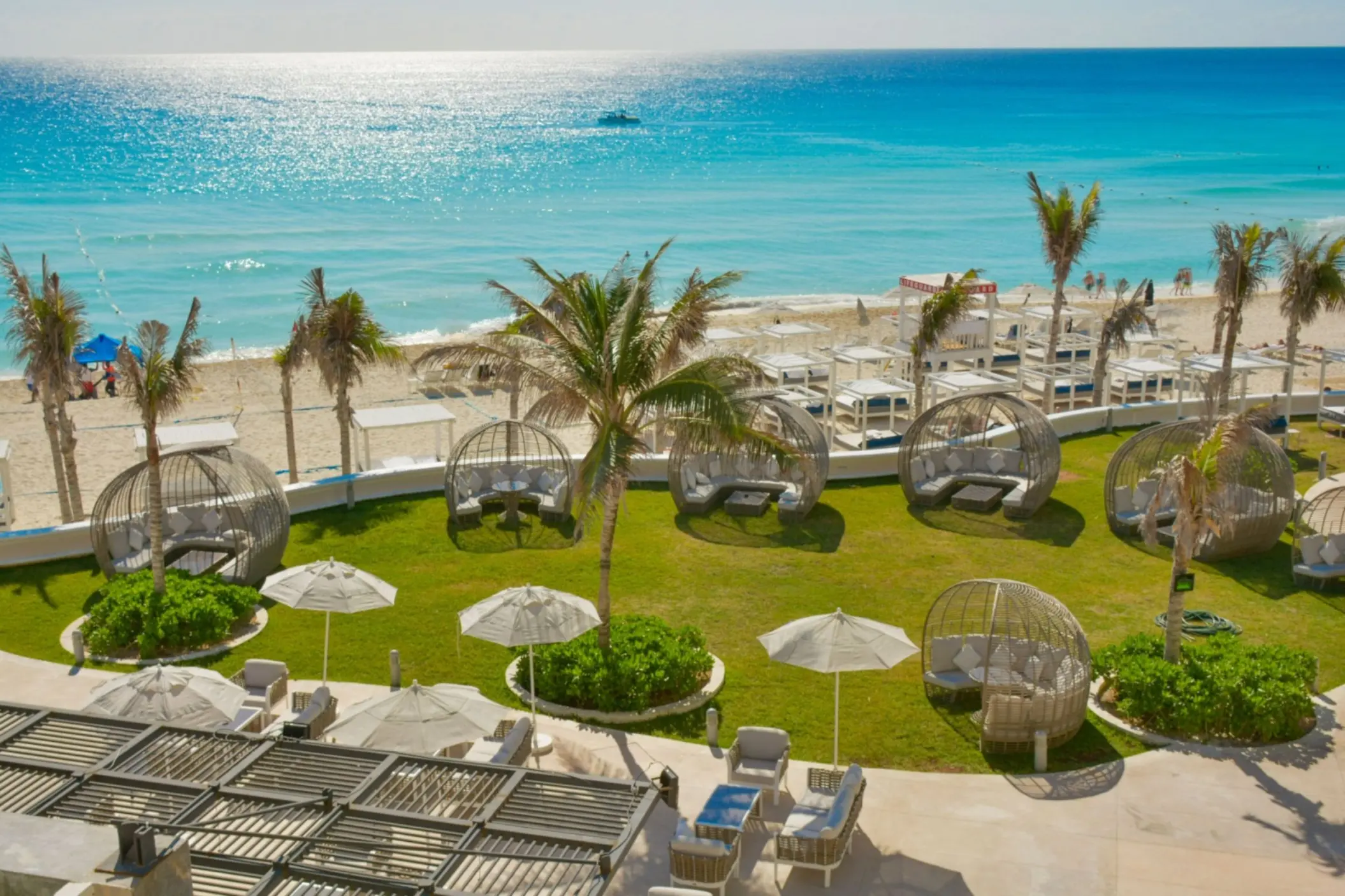 Aerial view of a grassy outdoor lounge area with palm trees, cabanas, and sun loungers near the beach.
