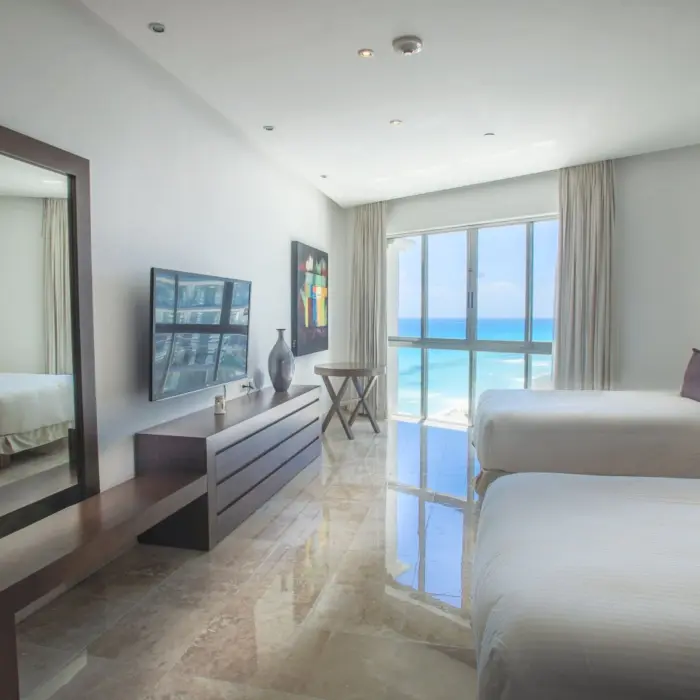 A bright, modern suite with two beds, a large mirror, and a wall of windows overlooking the ocean.