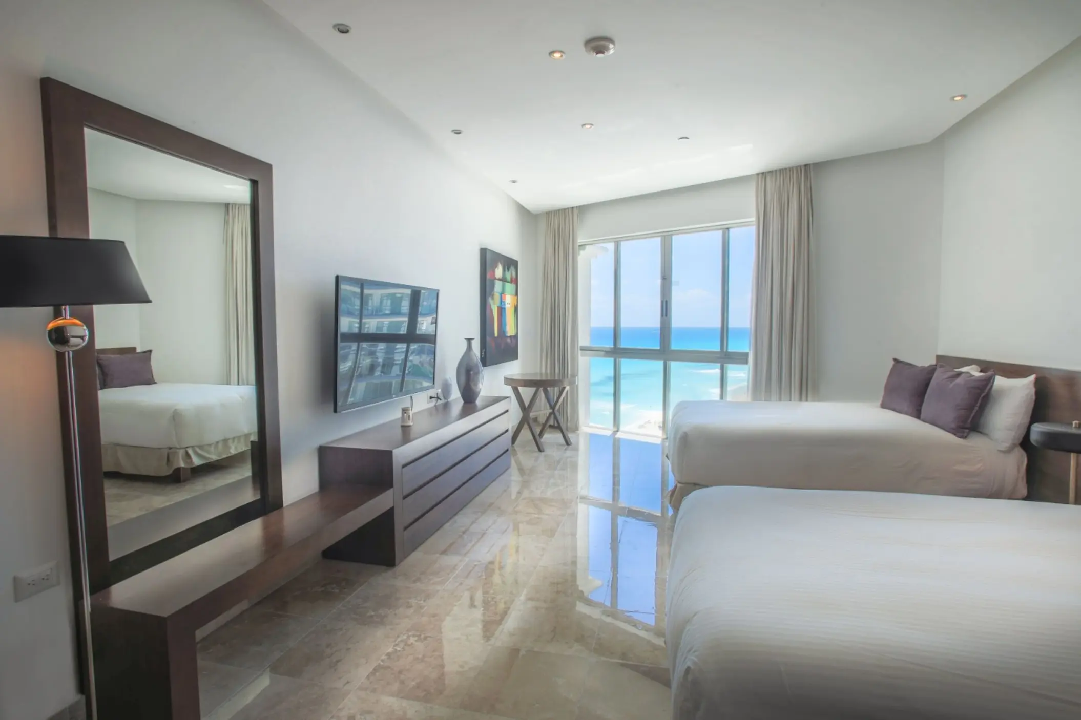 A bright, modern suite with two beds, a large mirror, and a wall of windows overlooking the ocean.