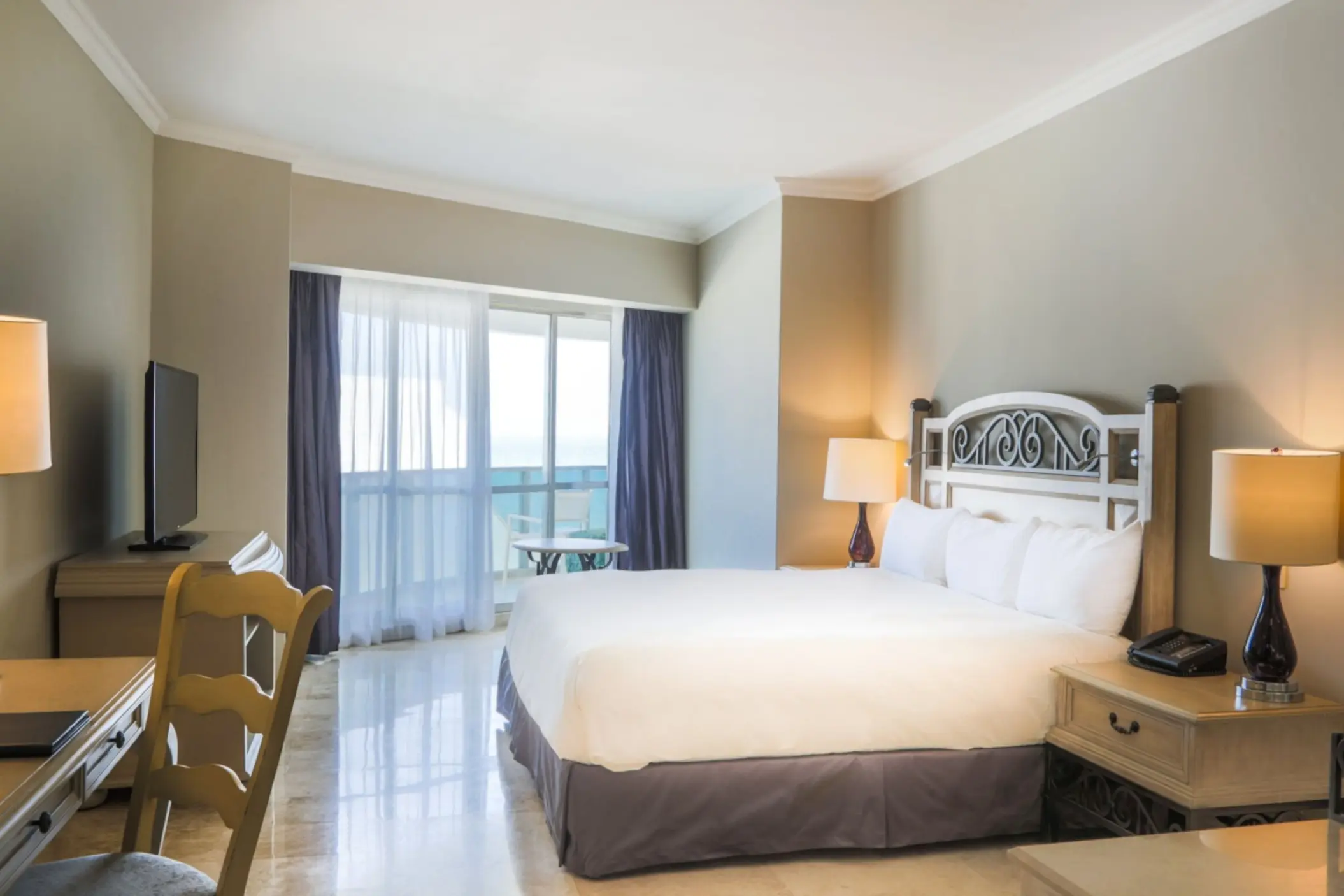 A spacious hotel suite with a king-sized bed, marble floors, a desk, and balcony access to a sea view.